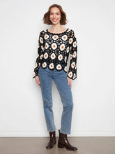 Load image into Gallery viewer, A woman modeling a black long-sleeve crochet top with a white and yellow daisy floral pattern, paired with blue jeans and dark brown ankle boots, standing against a white wall.

