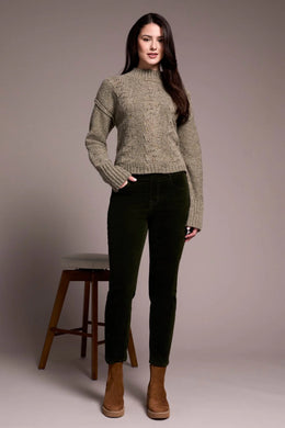 Woman wearing a green sweater and deep, dark green cord pants with brown boots standing next to a barstool against a plain background.