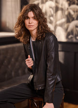 Load image into Gallery viewer, Woman wearing a black leather jacket, white tee and black pants, sitting on a chair against a blurred background.
