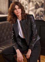 Load image into Gallery viewer, Woman wearing a black leather jacket, white tee and black pants, sitting on a chair against a blurred background.
