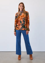 Load image into Gallery viewer, Woman wearing a leaf patterned blouse, blue jeans and camel-colored boots against a neutral background.
