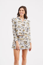 Load image into Gallery viewer, A woman modeling a matching long-sleeve collared top and skort set with a white base and gold and black graphic print, set against a plain light gray background.

