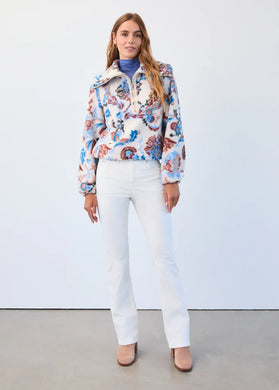 Woman wearing a paisley jacket over a blue turtleneck, creme colored corduroy denim pants and tan suede boots against a white wall.