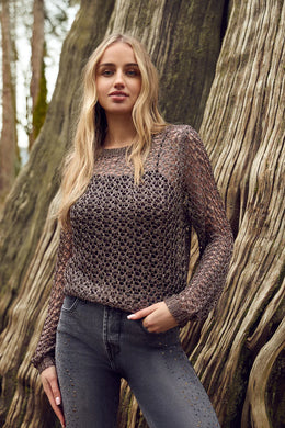 Woman wearing a sequined open knit sweater and jeans standing against a large tree trunk.