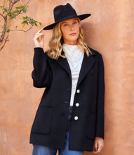 Load image into Gallery viewer, Woman wearing a black hat, black blazer with rhinestone buttons over a white top and blue jeans against an orange wall.
