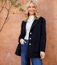 Load image into Gallery viewer, Woman wearing a black blazer with rhinestone buttons over a white top and blue jeans against an orange wall.
