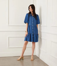 Load image into Gallery viewer, Woman wearing a blue chambray dress and sandals standing against a white paneled wall.
