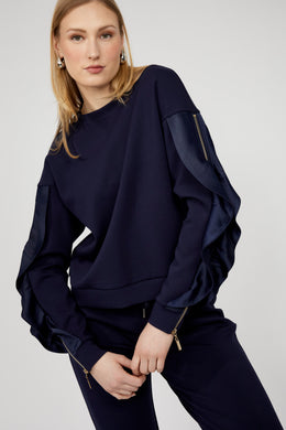 Woman wearing a navy blue ruffled sleeve with zippers top and navy blue pants against a white background.