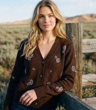 Load image into Gallery viewer, A woman wearing a brown knit cardigan with rhinestone floral embroidery, leaning against a rustic wooden fence in an outdoor desert landscape.
