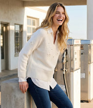 Load image into Gallery viewer, A woman laughing outdoors near a building and gas pumps, wearing a cream layer knit sweater with dark blue jeans.

