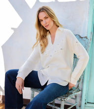 Load image into Gallery viewer, A woman sits on a distressed blue chair wearing a cream layered knit sweater with embroidered details, paired with dark blue jeans, against a white stucco wall.
