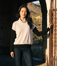 Load image into Gallery viewer, A woman wearing a color-blocked knit  turtleneck top with a cream body and black sleeves, paired with dark blue jeans, standing in a rustic stone archway with a desert landscape in the background.
