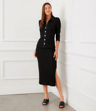 Load image into Gallery viewer, Woman wearing a black ribbed long sleeve top with buttons and long black skirt and black sandals standing against a white wall. 
