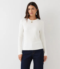 Load image into Gallery viewer, A woman modeling a white ribbed long-sleeve knit top with dark blue jeans, standing against a plain white studio background.
