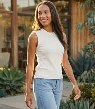 Load image into Gallery viewer, Woman wearing an off-white sleeveless tank op and blue jeans standing outdoors with plants and a wooden structure in the background.
