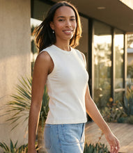 Load image into Gallery viewer, Woman wearing an off-white sleeveless tank top and blue jeans standing outdoors with a modern house in the background.
