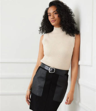 Woman wearing a cream sleeveless turtleneck sweater, black skirt and black belt against a white wall.