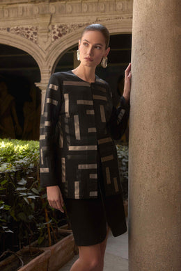 Woman wearing a patterned long jacket in black/multi and black skirt in an outdoor setting with architectural elements.