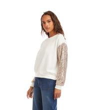 Load image into Gallery viewer, Woman wearing an off-white top with sequin sleeves and blue jeans against a white background.

