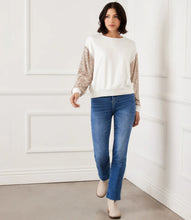 Load image into Gallery viewer, Woman wearing an off-white top with sequin sleeves, blue jeans and off-white boots against a white wall.
