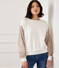 Load image into Gallery viewer, Woman wearing an off-white top with sequin sleeves and blue jeans against a white wall.
