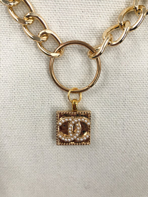 Introducing the extraordinary Shelly necklace! This piece features a rare, gold square adorned with a delicate pearl CC logo. The vintage Chanel button is elegantly suspended from a round connector, attached to a link chain. This exquisite necklace effortlessly complements any outfit. Get ready for endless compliments!