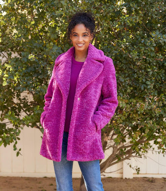 Woman wearing a bright orchid coat and blue jeans standing in front of a green tree.