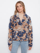 Load image into Gallery viewer, A woman modeling a long-sleeve button-up coat with a camel, navy, and white abstract floral pattern, paired with light-wash blue jeans and black ankle boots, standing against a white wall.

