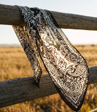 Load image into Gallery viewer, A sheer black and white paisley bandana scarf draped over rustic wooden fence rails in an outdoor field at sunset.
