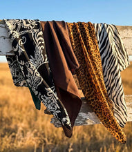 Load image into Gallery viewer, Four patterned fabric scarves  draped over a weathered wooden fence outdoors, with a field and blue sky in the background. The fabrics show black-and-white paisley, solid brown, leopard print, and zebra print patterns.
