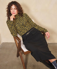 Load image into Gallery viewer, Woman sitting on a chair wearing a patterned top, long black skirt and black boots.
