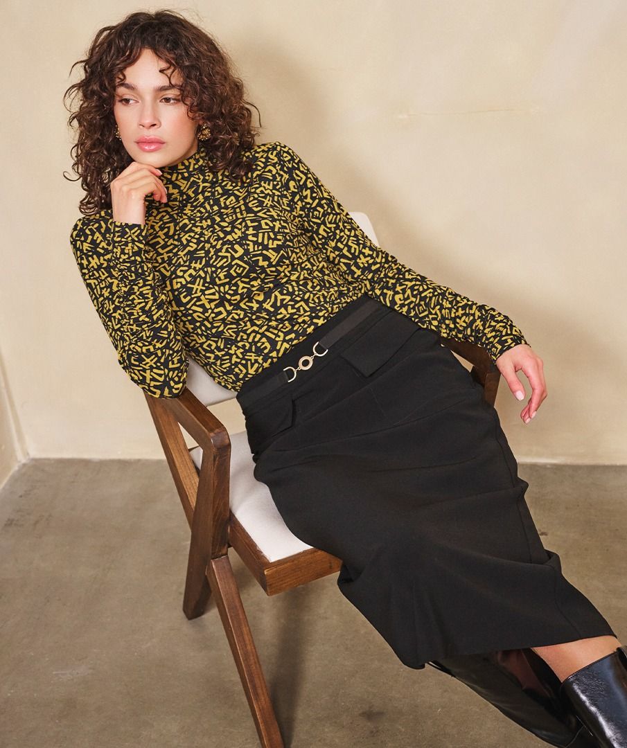 Woman sitting on a chair wearing a patterned top, long black skirt and black boots.