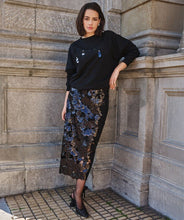 Load image into Gallery viewer, Model wearing a black sweatshirt, black sequined long skirt and black heels standing against a stone wall.
