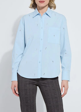 Woman wearing a light blue shirt with embellishments and dark plaid pants against a white background.