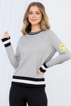 Load image into Gallery viewer, The Heather Gray Smiley Face Knit Sweater - TMK1517 from THML features classic black and white stripes on a gray background but adds a cheerful touch with a darling yellow smiley face on the upper part of each sleeve. This lightweight sweater has a vintage feel and modern appeal, with striping at the bottom hem, cuffs, and neck.  Color- Gray, black, white and yellow. Smiley face on upper sleeves. Black and white striping on neckline, sleeves and bottom hem. Lightweight sweater.
