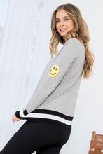 Load image into Gallery viewer, The Heather Gray Smiley Face Knit Sweater - TMK1517 from THML features classic black and white stripes on a gray background but adds a cheerful touch with a darling yellow smiley face on the upper part of each sleeve. This lightweight sweater has a vintage feel and modern appeal, with striping at the bottom hem, cuffs, and neck.  Color- Gray, black, white and yellow. Smiley face on upper sleeves. Black and white striping on neckline, sleeves and bottom hem. Lightweight sweater.
