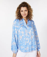 Load image into Gallery viewer, Our Sassa blouse with smock details on the chest and an all-over floral print on a striking blue background is one of a kind! This gorgeous blouse has a wider fit, a collar and button-through closure, while the sleeves have elasticated cuffs.   Colors- Blue, white and gray. Button front. Smocked design on chest. Wider fit. Elasticized cuffs.
