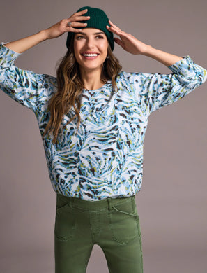 Woman wearing a green stocking cap, patterned green, blue, white sweater and green pants against a pale pink background.