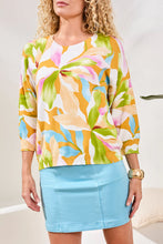 Load image into Gallery viewer, Woman in a colorful floral lightweight sweater and aqua skort standing in a bright room with a plant. 
