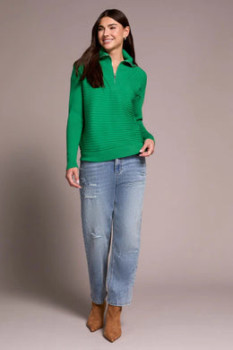 Woman wearing a green sweater, blue jeans and brown boots against a plain background.