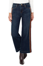 Load image into Gallery viewer, Model wearing dark blue wide leg jean with brown multicolor striping on sides, white long sleeve top and brown boots against a white background.
