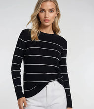 Load image into Gallery viewer, Woman wearing a black sweater with white stripes and white pants against a white background.
