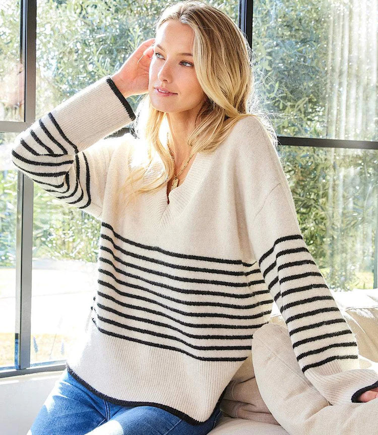 Woman wearing an ecru sweater with black stripes and jeans sitting indoors with large windows in the background.