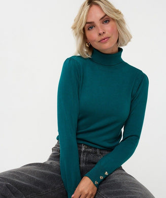 Woman wearing gold earrings, a teal turtleneck sweater with gold heart cufflinks and black jeans against a white background.