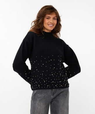 Woman wearing a black sweater with  black pearls on lower half of sweater and sleeves and grey pants against a white background.