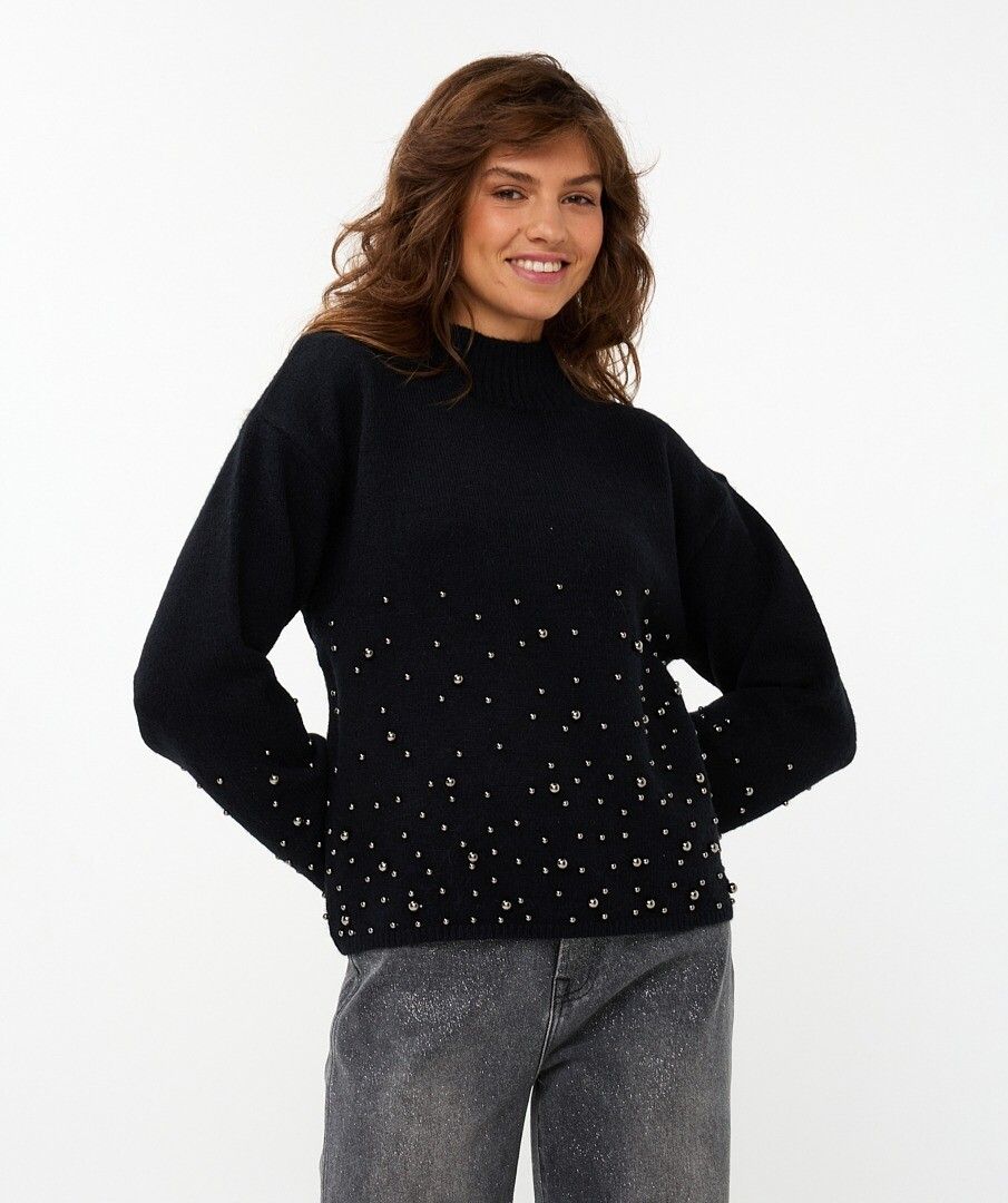 Woman wearing a black sweater with  black pearls on lower half of sweater and sleeves and grey pants against a white background.