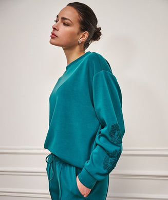 Woman wearing a teal modal sweater with floral embroidery and teal pants against a white wall background.