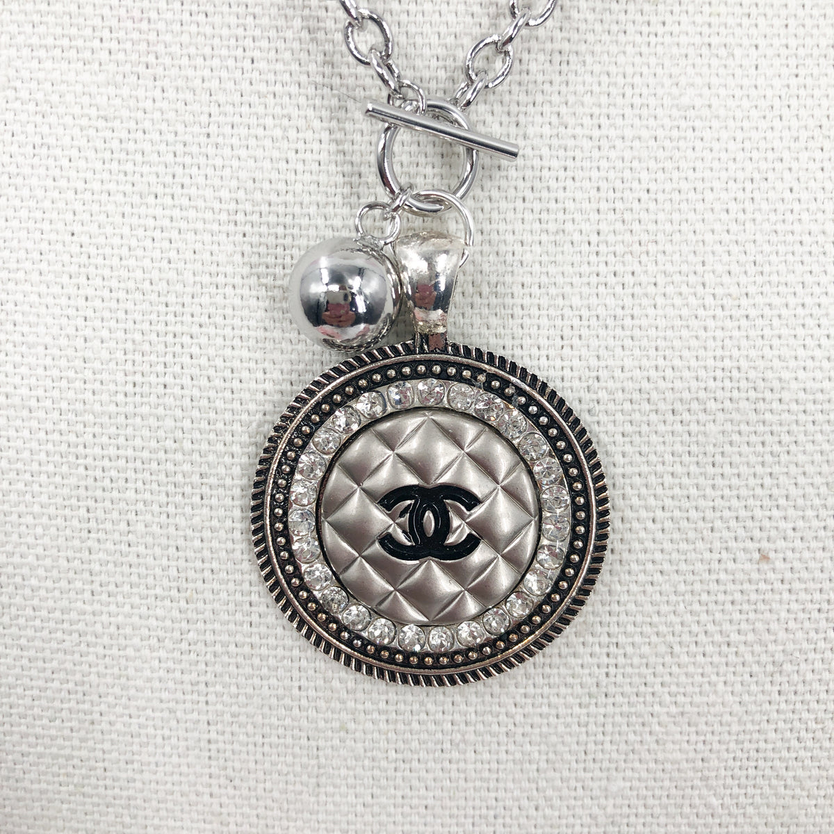 This necklace showcases a stunning and very rare silver vintage quilted button, embellished with a black CC logo. The exquisite button sits in a circular silver setting, encircled by brilliant clear rhinestones, and is suspended from a 925-sterling silver link chain that fastens with a toggle closure. Its unique allure is bound to garner an array of compliments.