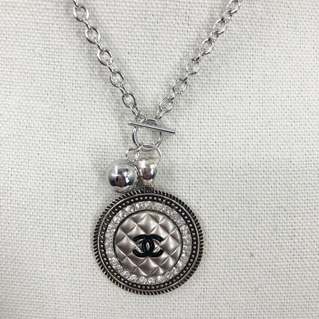 This necklace showcases a stunning and very rare silver vintage quilted button, embellished with a black CC logo. The exquisite button sits in a circular silver setting, encircled by brilliant clear rhinestones, and is suspended from a 925-sterling silver link chain that fastens with a toggle closure. Its unique allure is bound to garner an array of compliments.