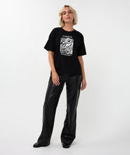 Load image into Gallery viewer, Woman wearing a black t-shirt with a graphic design, black pants and black shoes against a white background.
